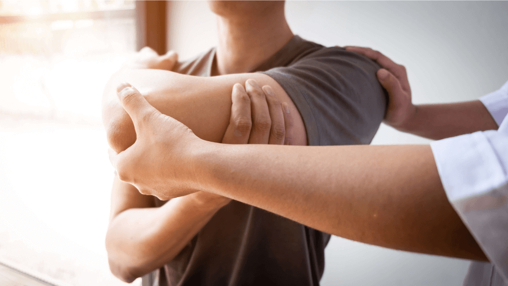 A person is receiving physical therapy as a therapist supports and stretches their arm near a window.