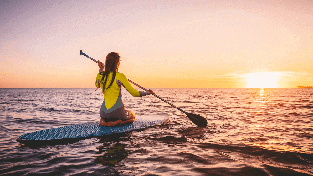 A person kneels on a Pro Balance 360 paddleboard, paddling on the ocean during sunset under a clear sky.
